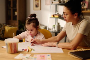 Genç anne ve Down sendromlu küçük tatlı kızı masa başında otururken kağıt üzerinde fosforlu kalemler çiziyorlar.