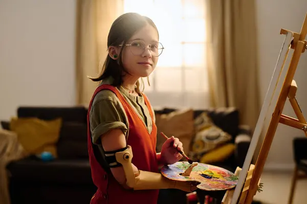 Pre-teen girl with disability looking at camera while standing in front of easel in living room and working over new creative artwork
