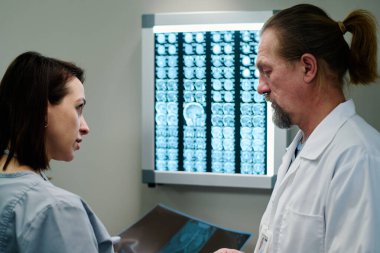 İki kendine güvenen klinisyen, muayene sırasında duvardaki genç asistan ve beyin taramasında röntgen görüntülerini tartışıyor.