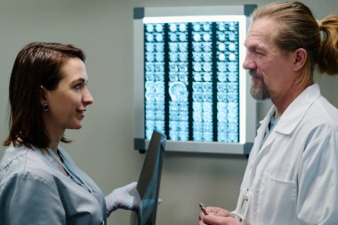 Röntgen görüntüsü olan genç bir radyolog ya da onkolog asistanı olgun bir doktora bakıyor ve ona sorular soruyor.