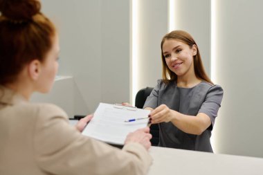 Üniformalı genç resepsiyonist ya da klinisyen tezgahın önünde duran kadın hastaya tıbbi belge veriyor.