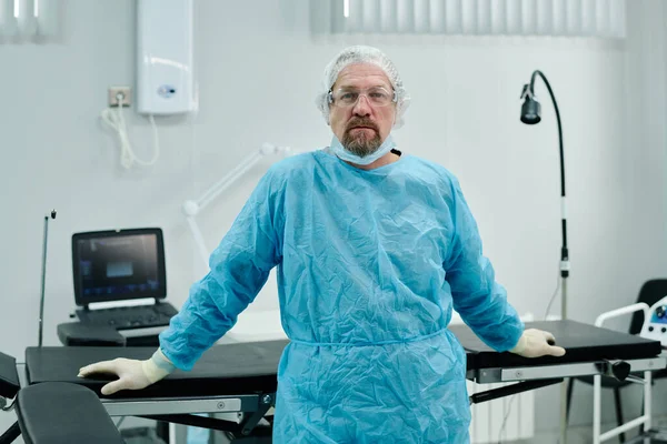 有丰富经验的外科医生 身穿蓝色的医疗服 戴着防护帽 面罩和眼镜 在他工作的地方看着摄像机 — 图库照片