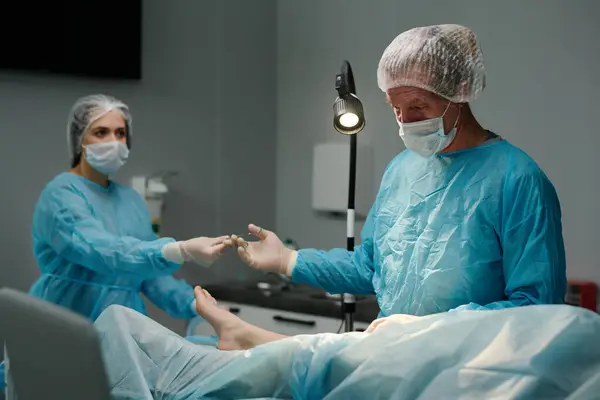 Mature male clinician taking sterile forceps from hand of gloved assistant in medical scrubs, cap and mask during surgical operation