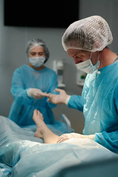 外科医生在手术过程中从女助手手里接过医疗工具 带着医疗刷子 帽子和面罩 — 图库照片