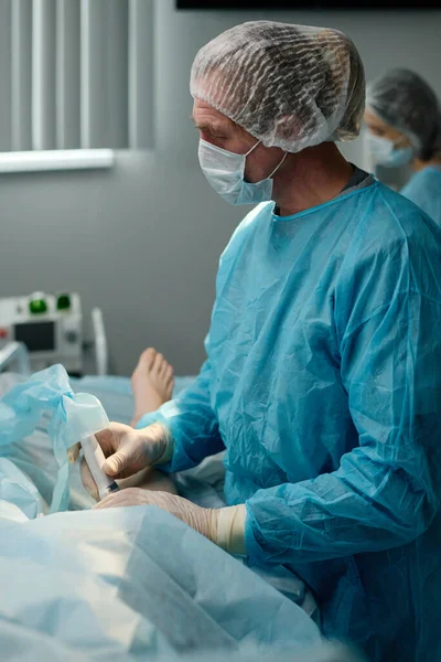 Experienced male clinician with probe standing by operating table with patient covered with napkin during surgical operation