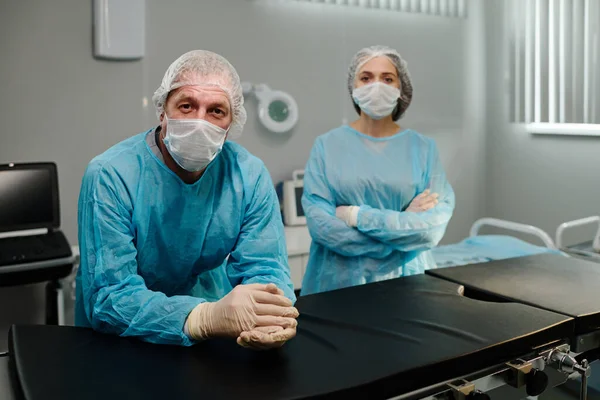 Mature male surgeon and young female assistant in medical scrubs, caps, masks and gloves looking at camera while standing by operating table