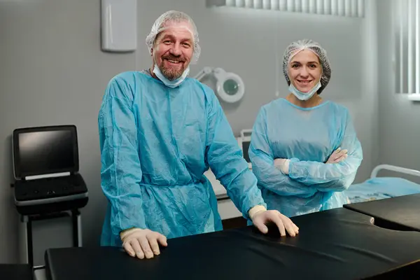 ハッピー外科医と医療スクラブのアシスタント 保護キャップ マスク 手袋は オペレーティングルームの職場で立っています — ストック写真