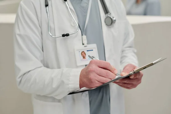 Ręka Męskiego Lekarza Stetoskopem Odznaką Notuje Dokumencie Stojąc Przed Kamerą — Zdjęcie stockowe