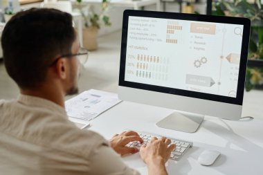 Genç erkek İK yöneticisi klavyede yazı yazıyor ve ofiste çalışırken bilgisayar ekranındaki verilere bakıyor