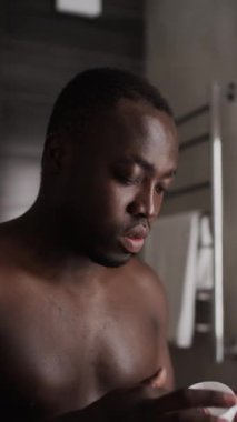 Genç Afrikalı Amerikalı adamın, kavanozdan bir tutam nemlendirici yüz kremi alıp yanaklarına sürerken, aynaya bakarken, günlük cilt bakımı sırasında