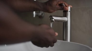 Evdeki beyaz seramik havzada banyo musluğundan musluk kaldıran ve akan suyun altında ellerini yıkayan tanınmayacak kadar genç siyahi veya Asyalı bir adamın çıplak elleriyle yakın çekim.