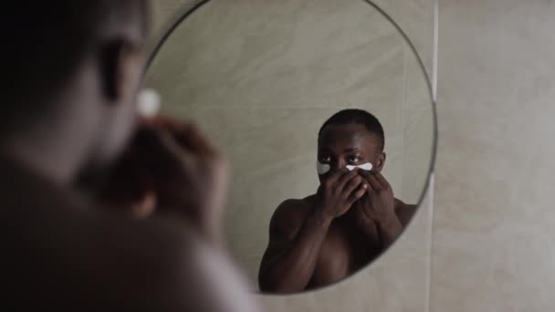 年轻的非洲裔美国男子 胸部裸露 在眼罩下小心涂上润肤液 同时在浴室镜子中看到自己的倒影 — 图库视频影像