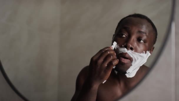 年轻的非洲裔美国人 光着胸站在浴室镜子前 用一次性剃须刀精心刮胡子 看着自己的倒影 近照肩膀 — 图库视频影像