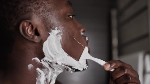 自宅で使い捨てカミソリと白い泡の上に髪剃りとひげを切った若いアフリカ系アメリカ人男性のクローズアップサイドショット — ストック動画