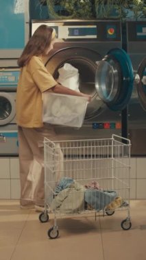 Kafkasyalı genç bir kadının plastik sepet taşıdığı ve çamaşırhanedeki çamaşır makinesine beyazı yüklediği dikey yan görüntüler.