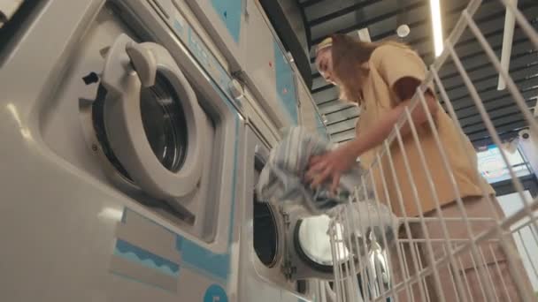 セルフサービスの公共ランドリーで洗濯機にカートから汚れた服をロードするコーカサス州の若い女性の低い角度の映像 — ストック動画