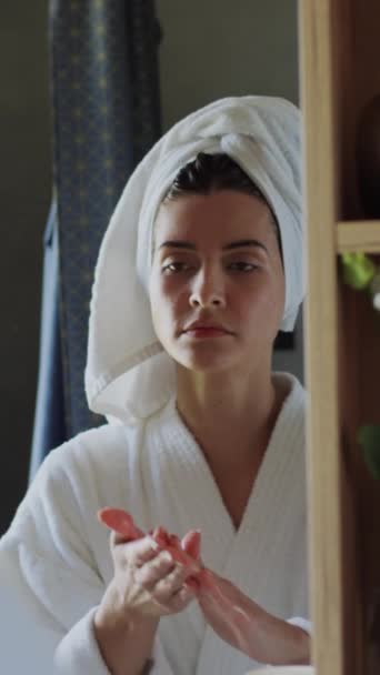 白种人或西班牙裔妇女在浴袍中的垂直特写 毛巾头巾中的湿头发 从瓶子中取出乳液 在手掌间揉搓 照镜子时在脸上撒布 — 图库视频影像