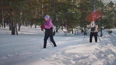 Yünlü şapkalı yaşlı kadın mercekli adam ve anorak ceketli adam, huş ağacında ve çam ormanlarında kayak yaparken soğuk güneşli bir günde kar altında kameraya yakalandılar.