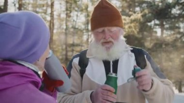 Kış günü orman yürüyüşü sırasında vakum şişelerinden gelen buharlı sıcak çayın keyfini çıkarırken, kalın gri sakallı, eşiyle sohbet eden yaşlı, beyaz bir adamın lens ışığıyla orta boy fotoğraf.