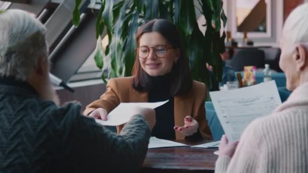 カフェで高齢者カップルとのビジネスミーティングを行っている女性金融コンサルタントの中間ショット 退職年金プランを提示し 署名するための法的文書のコピーを与え 丁寧に微笑む — ストック動画