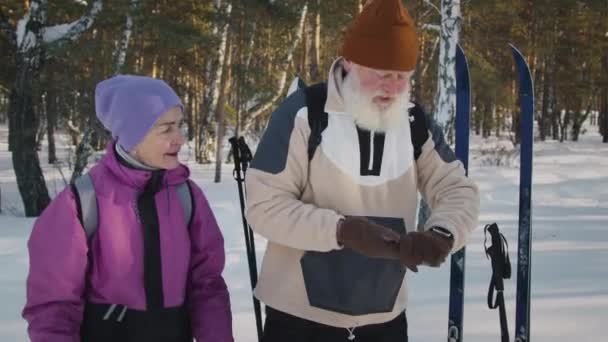 ビーニーと暖かいジャケットで高齢の白人妻と 冬の森でのスキー旅行中に時計を見て 笑顔で笑い お互いに高揚感を与え合った夫のメディアショット — ストック動画