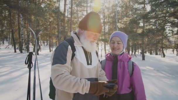 中镜头闪烁着白种人快乐的镜头 白种人留着浓密的灰胡子 头戴羊毛帽 妻子一边用智能手机自拍 一边看冬日滑雪时的照片 — 图库视频影像