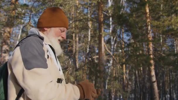 厚い灰色のひげを持つ成熟した白人男性のミディアムアークショット 暖かいアナマで バックパックは手袋に入れ ウリー帽子を引っ張り 森で一人でハイキングする前にスキーポールを取ります — ストック動画