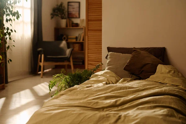 Part of spacious empty bedroom with comfortable undone bed with two cushions standing in the corner of room with green plants