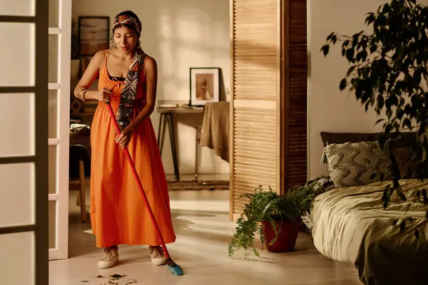 Saç bandı takmış genç bir İspanyol kadın ve hafta sonları ev işleri yaparken yatak odasının yerlerini fırçayla temizliyor.