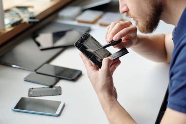 Küçük tornavidayla tamir merkezinde çalışan genç işçinin elleri iş yerindeki parçalanmış cep telefonunun küçük detaylarını onarıyor.