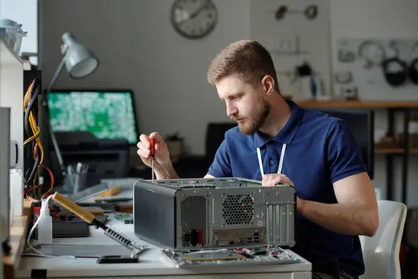 修理サービスオフィスの職場とコンピュータプロセッサの固定部分に座ってダークブルーのユニフォームの若い深刻な男性技術者 — ストック写真