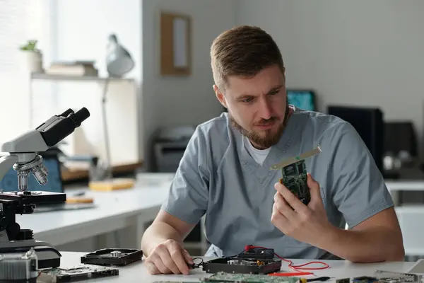 職場に座って技術的な問題を特定しながら コンピュータプロセッサの一部を見ている灰色のユニフォームの若い深刻な男 — ストック写真