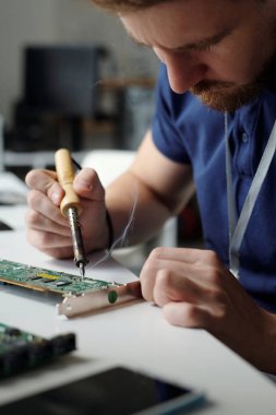 Bakım işlerinden sorumlu genç erkek mühendis kameranın önünde oturuyor ve bilgisayar anakartını elektrikli el aletleriyle onarıyor.