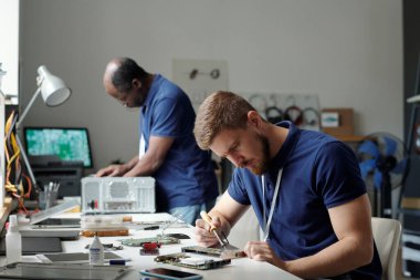 Genç ciddi sorun çözme ve tamir servisi uzmanı şanzıman cımbızla anakart ile iş yerinin üzerine eğiliyor.