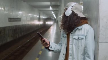 Saçları kıvırcık, kulaklıkla müzik dinleyen ve metro istasyonunun durmasını beklerken cep telefonu ekranına bakan beyaz bir adamın yan görüntüleri.