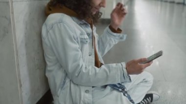 Kot ceketli ve kot pantolonlu beyaz bir adamın uzun ve boş bir yeraltı lobisinde oturup müzik dinlediği yan görüntüler.