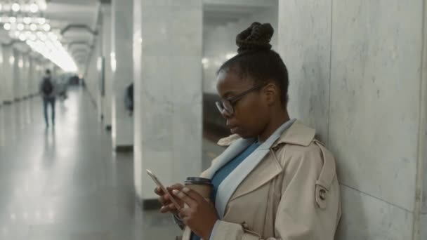 サイドチェストアップ映像 メガネでスタイリッシュな黒人女性は 電話でメッセージをチェックしながら 地下鉄駅で彼女の列車を待って 使い捨てコーヒーカップ — ストック動画