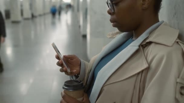 地下鉄の廊下で地下鉄を待っている間 モバイルと使い捨てカップからコーヒーを読み取るトレンチコートのアフリカ系アメリカ人女性の側傾きのクローズアップ — ストック動画