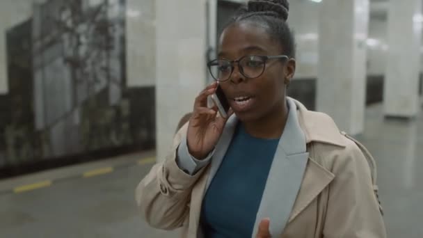 メガネとトレンチコートの陽気なアフリカ系アメリカ人女性のサイドトラッキング映像は 空の地下鉄ロビーに沿って一人で歩き 電話で話す — ストック動画
