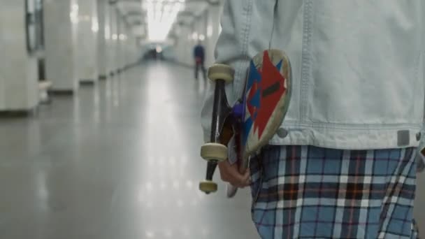 若いハンサムな白人カーリーヘアのスケートボーダーのチルトトラッキング映像は タイルの地下鉄の地下パスでロビーに沿って歩いている明るいボードで — ストック動画