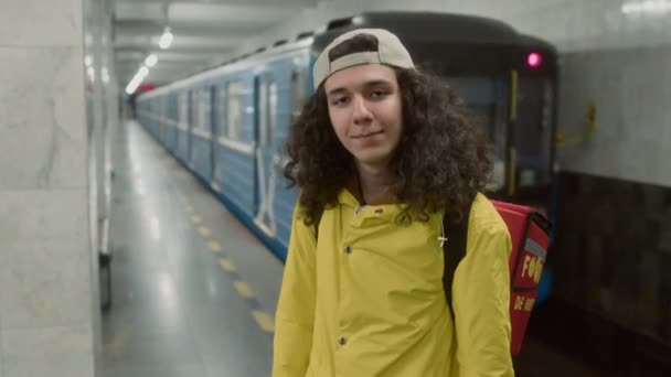 赤いバックパックを着た黄色いジャケットのハンサムなカーリー髪の男性宅配便の肖像画と地下鉄を離れることに対して立っているカメラを見る — ストック動画