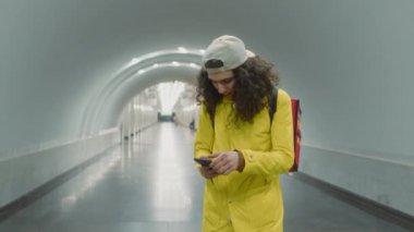 Sarı ceketli, kırmızı çantalı, akıllı telefondan adres kontrol eden ve metro istasyonuna giren genç teslimatçının geniş bir görüntüsü.