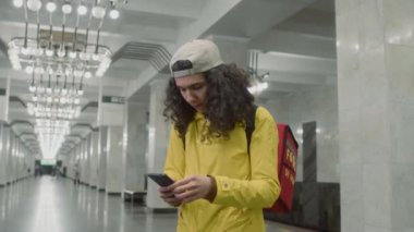 Sarı ceketli genç kuryenin yan görüntüleri. Metro koridorunda telefon kullanarak kırmızı sırt çantasıyla yemek dağıtırken.