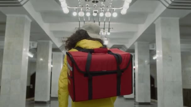 空の地下鉄廊下を歩いている大きな赤いバッグで食べ物を届ける未確認の宅配便の遅い追跡 — ストック動画