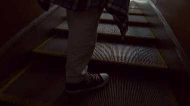 Arka tarafta, patenci tarzı giyinmiş, yürüyen merdivenden inip metro çıkışına giden kimliği belirsiz bir yolcunun görüntüsü var.