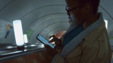 Trençkotlu Afrikalı Amerikalı kadının telefon ekranındaki metro transit haritasına bakarken yeraltı istasyonuna inen yürüyen merdivenin görüntülerini kaldır.