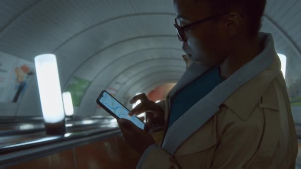 トレンチコートのアフリカ系アメリカ人女性の横の胸は 地下駅にエスカレーターを降下しながら 携帯電話の画面上の地下交通マップを見ています — ストック動画