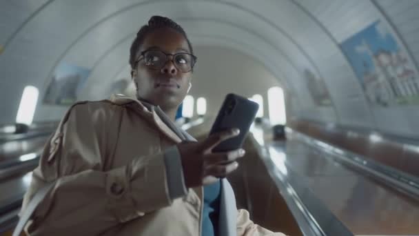 アフリカ系アメリカ人の女性通勤者がメガネをかけ 車内で電話を見ながら地下鉄の出口にエスカレーターを上昇させる様子 — ストック動画