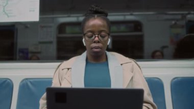 Gözlüklü ve kulaklıklı Afrikalı Amerikalı 'nın metroyla seyahat ederken dizüstü bilgisayarla uzaktan çalışan görüntülerine bak.