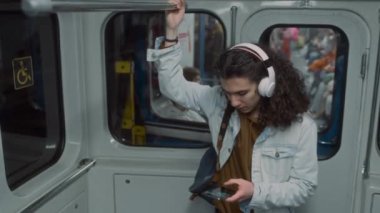 Kafkasyalı lise öğrencisi ya da üniversite öğrencisi metro treniyle seyahat ederken akıllı telefon kullanarak tırabzanlara tutunurken ve kulaklıkla ses dinlerken orta ölçekli görüntüler.